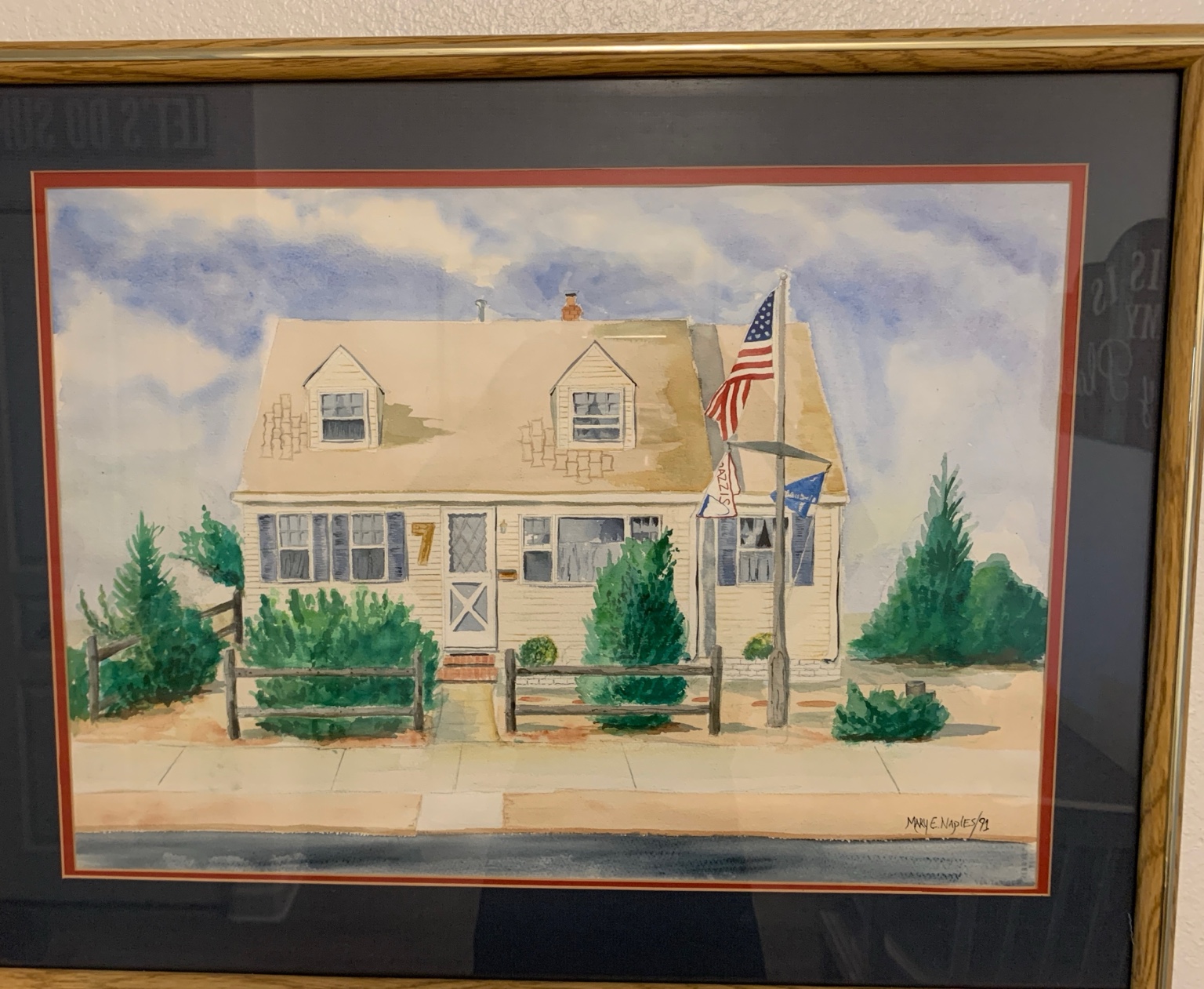 Framed watercolor painting of a seaside cottage with an American flag and greenery